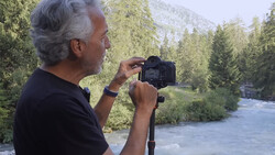 Photographer holding DSLR camera while composing a shot of a mountain river surrounded by evergreen forest.