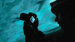 Person holding a camera with illuminated LCD screen against a blue-tinted background.