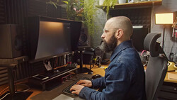 Bearded man working at a computer desk in a professional studio setup with monitor, keyboard, and audio equipment.