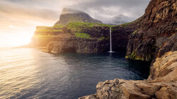 Waterfall cascading into a sea cave beneath towering coastal cliffs at golden hour.