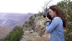 Woman with long brown hair photographing a vast canyon landscape from a rocky cliff overlook.