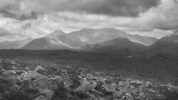 Mountain range with rocky foreground under dramatic cloudy sky in black and white.