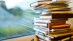 Stack of closed books and notebooks beside a window with blurred green landscape in background.