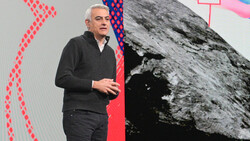 Speaker gesturing while presenting on stage with abstract geometric graphics and a black and white mountain photograph displayed on large screens behind him.