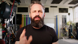 Bearded man in black t-shirt gesturing while speaking in a studio with professional lighting equipment visible.