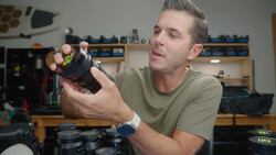 Man in tan shirt examining a black camera lens in a workshop setting.
