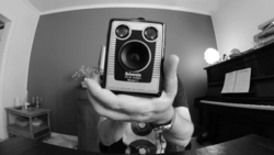 Photographer holds a vintage square-format camera with dual lenses toward the camera in a black and white self-portrait.