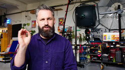 Bearded man in purple work shirt gesturing in a studio workshop filled with photography equipment and lighting gear.
