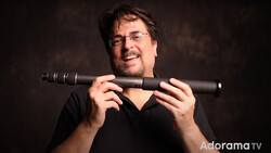 Man wearing glasses holding a telephoto lens toward the camera against a dark background.