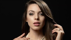 Woman with long dark hair wearing pearl necklace and ring, posed against black background.