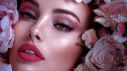 Woman's face surrounded by pink and mauve roses, with soft directional lighting and polished makeup.