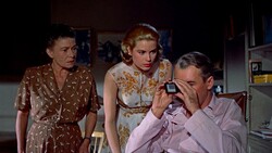 Three people in a 1950s interior setting, with a man holding a camera while two women observe.