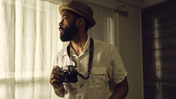 Photographer holding vintage film camera while looking out window with natural sidelight.