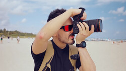 Photographer holding a DSLR camera with telephoto lens at the beach during daytime.