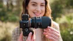 A photographer holds a mirrorless camera with an attached telephoto lens toward the camera, smiling outdoors.