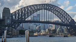 Steel arch bridge spanning a harbor with modern city skyline and waterfront buildings visible beneath it.
