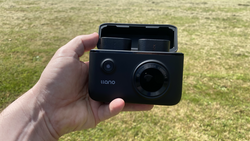 Hand holding an open Ilano compact camera against a grass background.