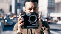 Photographer holding Canon EOS R5 camera body in urban setting with blurred cityscape background.