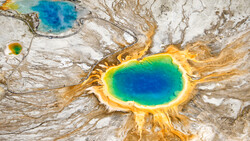 Aerial view of a geothermal hot spring with concentric rings of blue, green, and yellow mineral deposits.