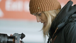 Female videographer with professional camera wearing tan beanie and black jacket.