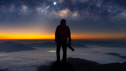 Silhouetted figure standing on a mountain peak overlooking clouds at sunrise with the Milky Way visible above.