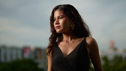 Woman with long dark wavy hair wearing black sleeveless top and gold earrings, photographed outdoors against blurred cityscape.
