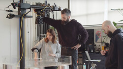 Three people collaborating around a 3D printer in a bright workshop studio setting.