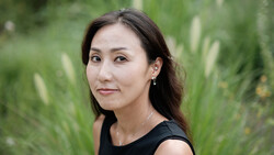 Woman with long dark hair wearing black sleeveless top, posed outdoors against soft green foliage background.