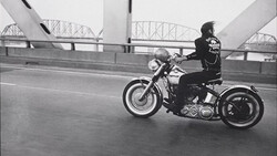 Motorcyclist riding a cruiser on a bridge with architectural steel framework visible in the background.