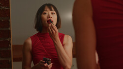 Woman in red sleeveless turtleneck applying lipstick while looking in mirror.