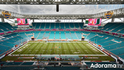 Large stadium with turquoise seats and green field viewed from upper seating level under steel roof structure.
