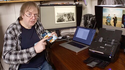 Photographer holding a light meter while seated at a desk with a laptop and scanner.
