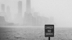 A "Danger No Swimming" sign stands in the foreground beside foggy water with a city skyline barely visible in heavy mist.