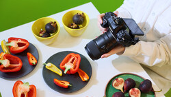 Photographer holding DSLR camera over styled food photography setup with fresh vegetables and fruits.