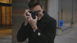 Young man in black jacket holding a rangefinder camera to his face, smiling while shooting in an urban corridor.