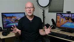Man in black shirt gesturing while seated at a studio desk with photography equipment and monitors displaying architectural images.