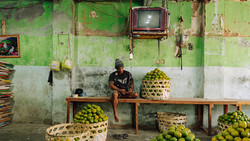 Man sitting on wooden bench in front of green deteriorated wall surrounded by baskets of limes.
