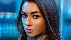 Woman with dark hair looking directly at camera against blue-tinted background.
