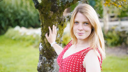Young woman with blonde hair wearing a red polka dot dress, leaning against a moss-covered tree in a garden setting.