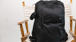 Black camera backpack displayed on a wooden chair with white canvas seat and back.