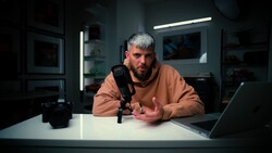 Man speaking into microphone at desk with laptop in dimly lit studio setup.