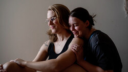 Two smiling people sitting together against a neutral gray background, looking downward in a candid moment.