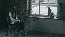 Person with long dark hair sitting on a wooden chair in a deteriorating room, gazing toward a large window overlooking water.