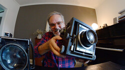 Photographer demonstrating a large format film camera held up to his face in an indoor studio setting.