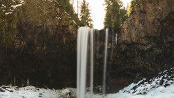 Waterfall cascading between tall rock cliffs surrounded by evergreen forest and snow.
