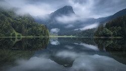 Mountain lake with forested shores and misty peaks reflected in calm water.