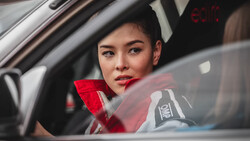 Woman in red jacket sitting in car's driver seat, looking toward camera through windshield.