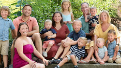 Multi-generational family group of 15 people posed together outdoors against green foliage.
