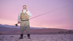 Fly fisherman in waders standing in shallow water at dusk with mountains in background.