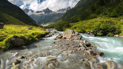 Turquoise river flowing through an alpine valley with snow-capped peaks and lush green meadows.